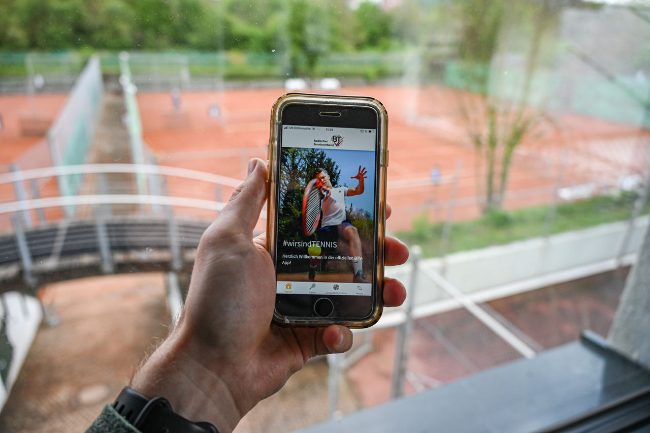 Hand mit Handy in der Hand und Tennisplätzen im Hintergrund